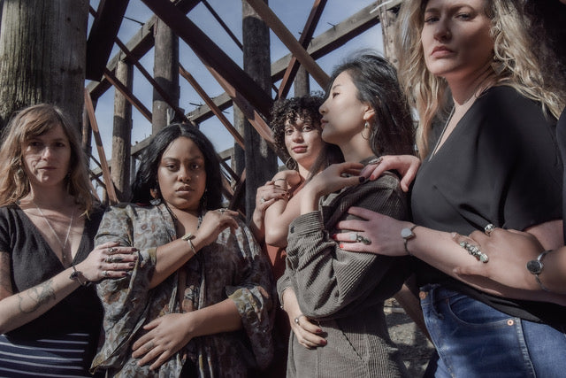 five different women standing on the banks of the Willamette River at an abandoned ship yards, modeling beautiful, handmade jewelry in a huddle.